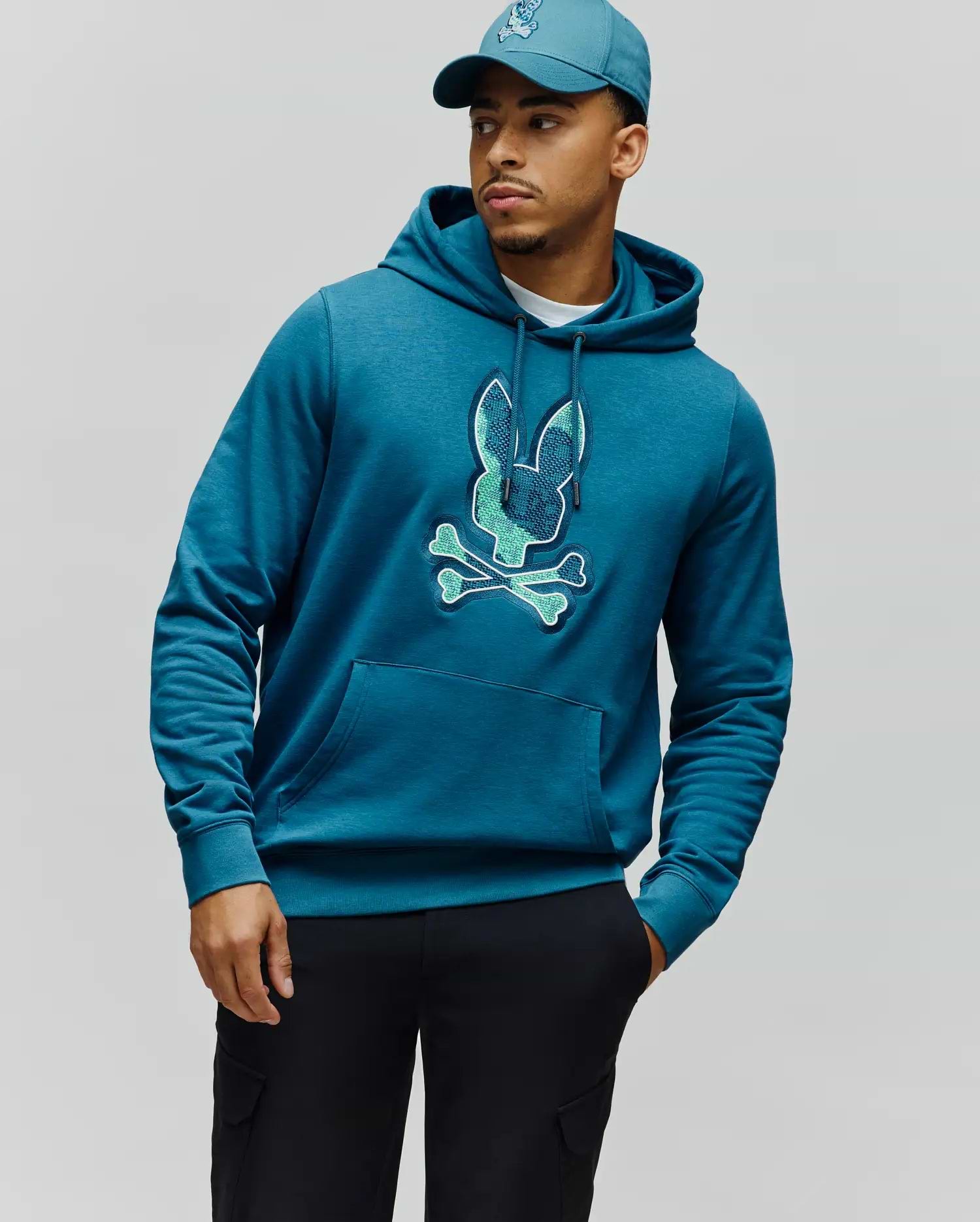Mens Olivier Embroidered Hoodie, a classic fit teal blue heathered jersey hoodie featuring a large, central embroidered Psycho Bunny skull logo in light teal and green, with a drawstring hood and kangaroo pocket. (3)