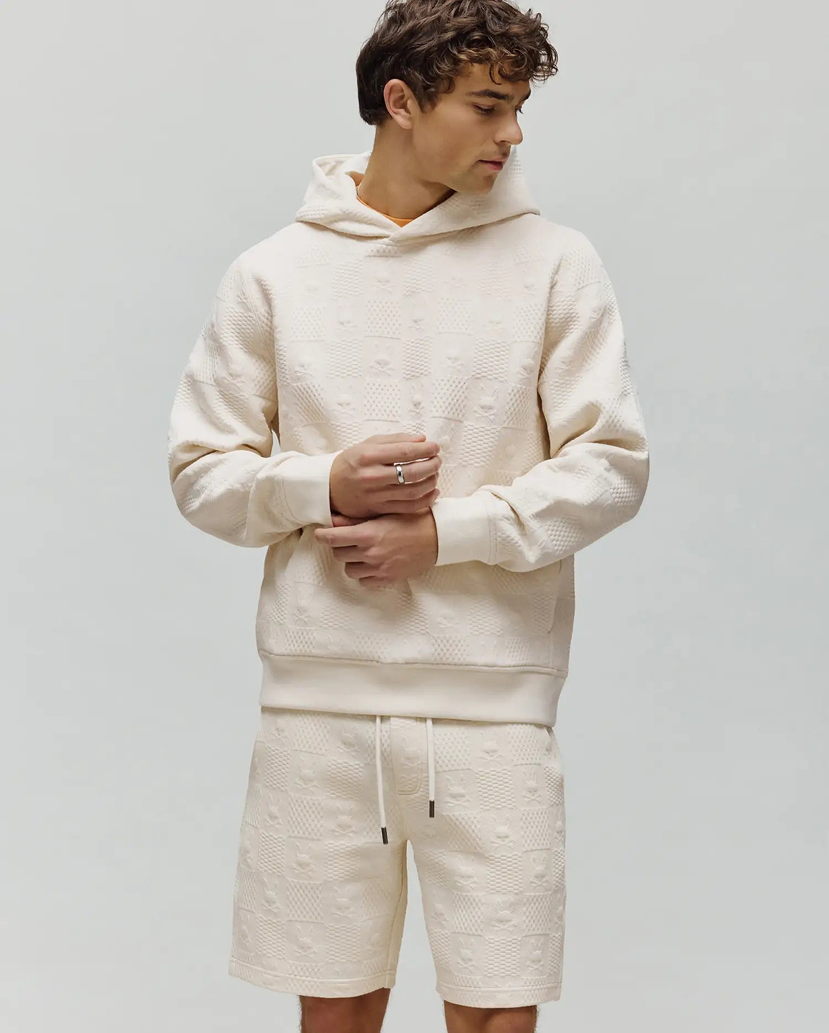 Mens Dustin Hoodie, a cream relaxed fit hoodie featuring an all-over embossed Psycho Bunny skull and crossbones logo pattern with a textured checkered design and a subtle orange neckline.