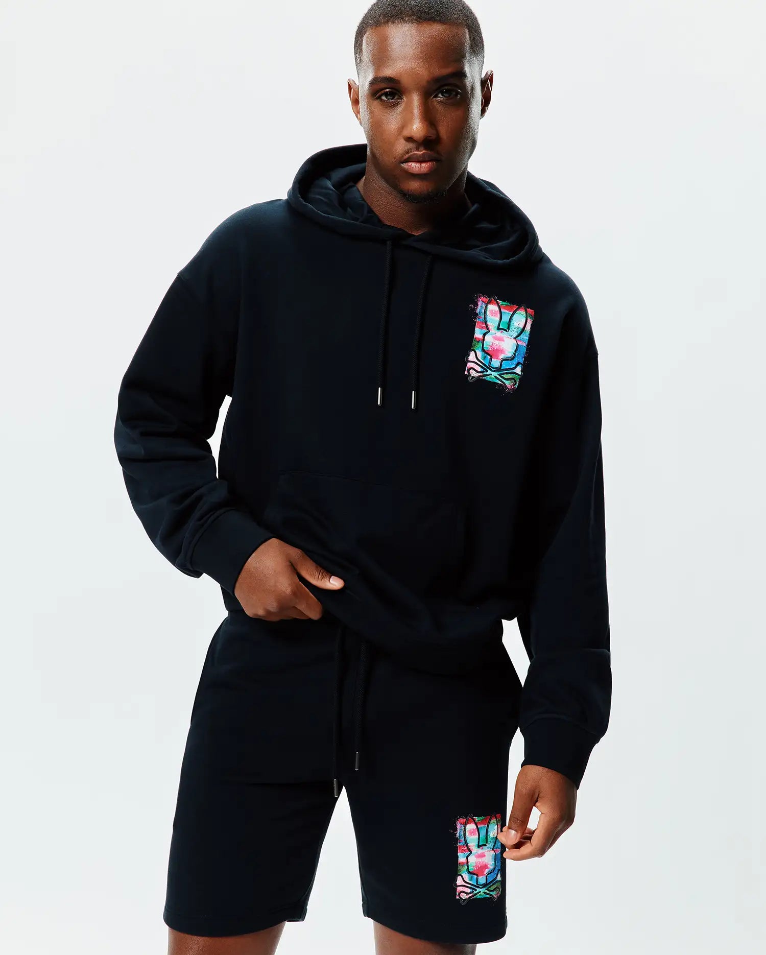 Soft black Mens Isaac Print Hoodie and matching shorts, featuring a relaxed fit. Both pieces showcase a large, vibrant, multi-color Psycho Bunny skull print with a distressed effect on the left chest and left leg.