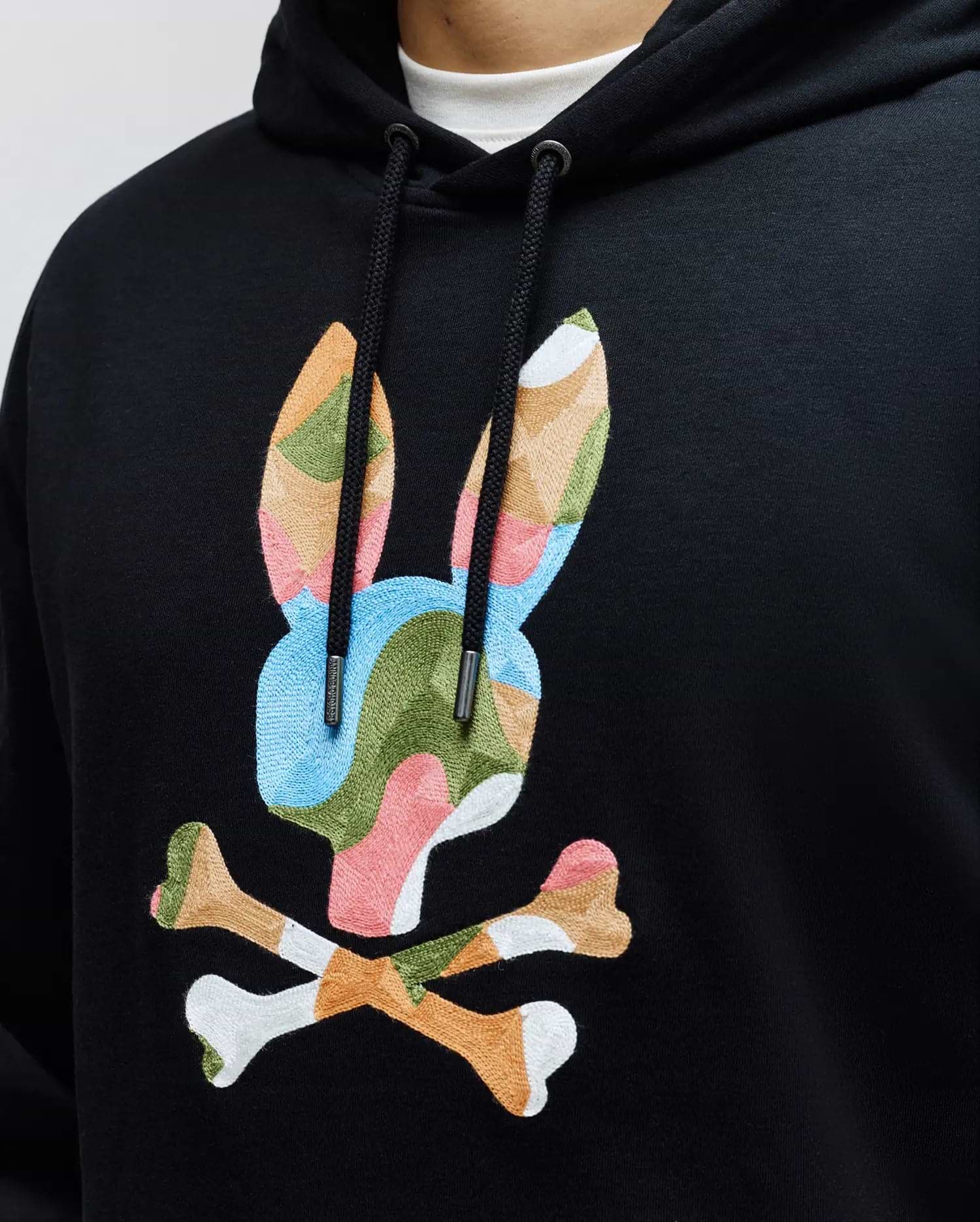 Mens Ara Hoodie: Black french terry hoodie with a large, central embroidered Psycho Bunny skull logo in a vibrant multi-color camouflage pattern, featuring branded drawstring tips and a relaxed fit.