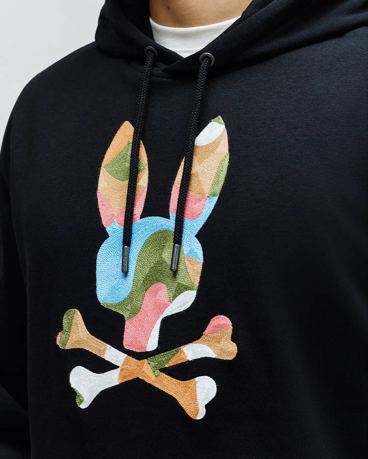 Mens Ara Hoodie: Black french terry hoodie with a large, central embroidered Psycho Bunny skull logo in a vibrant multi-color camouflage pattern, featuring branded drawstring tips and a relaxed fit.