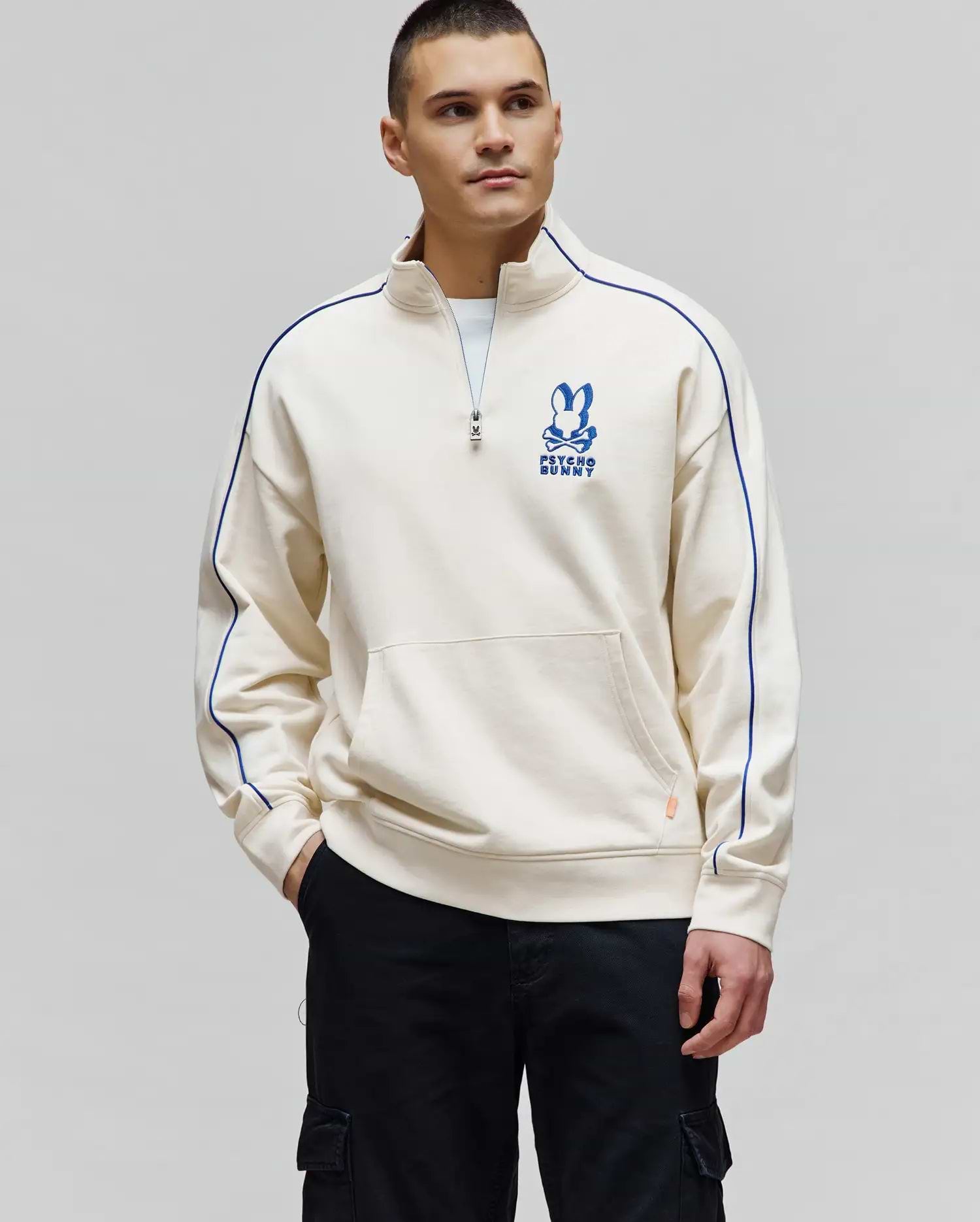 Mens Toby 1/4 Zip in cream French terry, featuring a relaxed fit, stand-up collar, royal blue piping on shoulders and sleeves, a large embroidered blue Psycho Bunny skull logo on the left chest, and a front kangaroo pocket. (1)