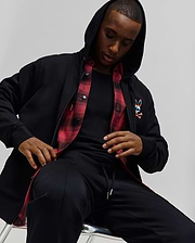 Mens Raphael Zip Hoodie in black, featuring a classic fit, smooth fabric, full zip, hood, ribbed cuffs, and a vibrant embroidered Psycho Bunny skull logo on the left chest, with a red plaid lining visible.