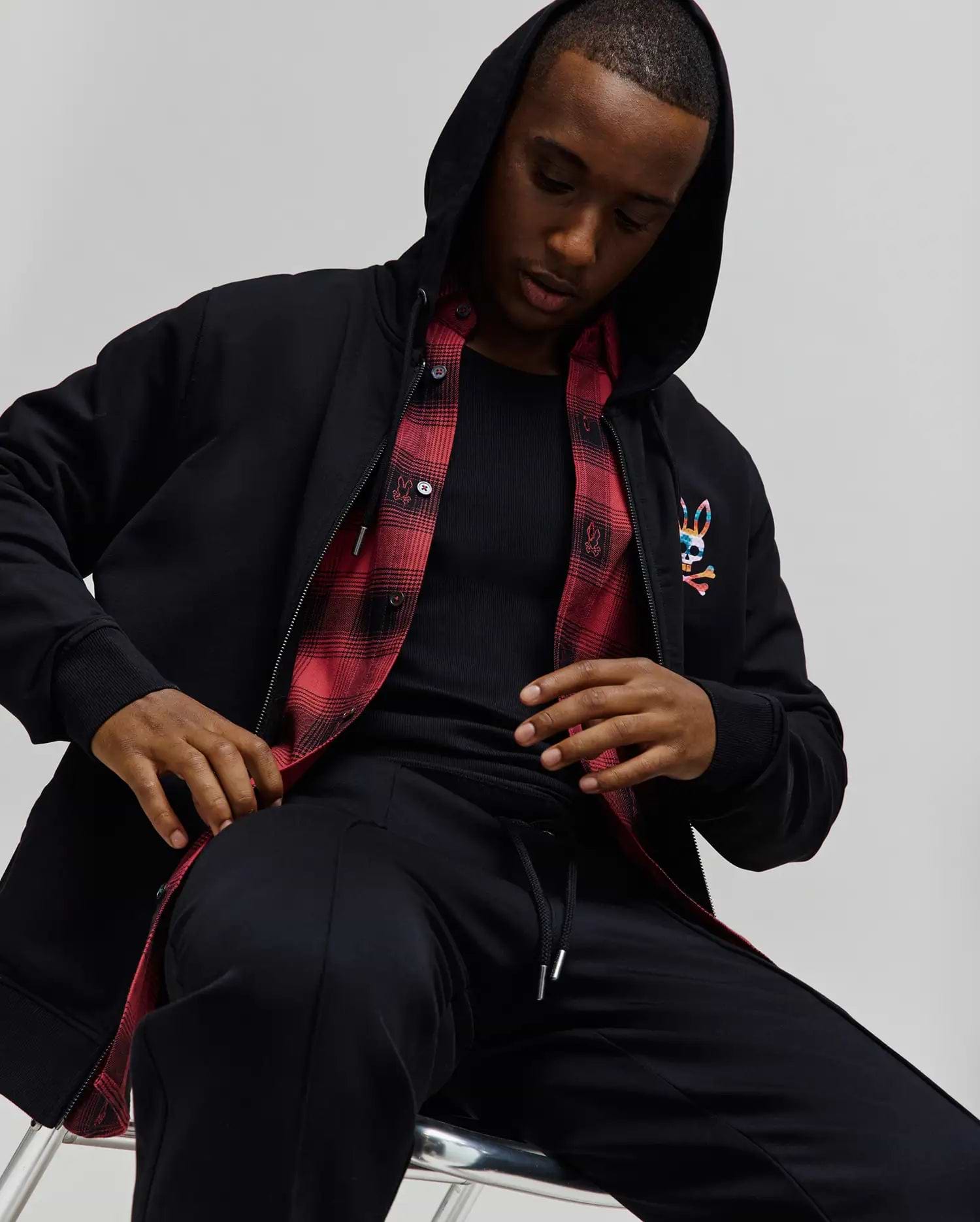 Mens Raphael Zip Hoodie in black, featuring a classic fit, smooth fabric, full zip, hood, ribbed cuffs, and a vibrant embroidered Psycho Bunny skull logo on the left chest, with a red plaid lining visible.
