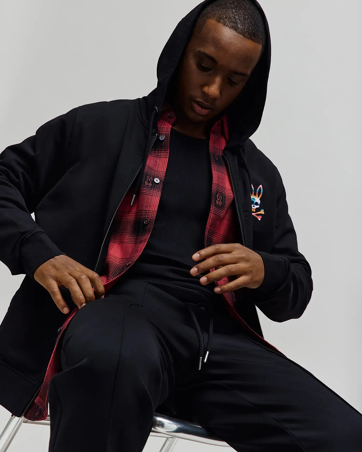 Mens Raphael Zip Hoodie in black, featuring a classic fit, smooth fabric, full zip, hood, ribbed cuffs, and a vibrant embroidered Psycho Bunny skull logo on the left chest, with a red plaid lining visible.