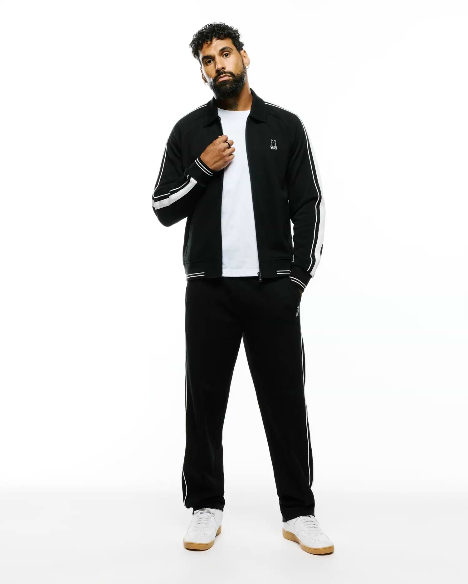 Black Mens Ceano Track Jacket and matching track pants, featuring white double sleeve stripes, single pant stripe, and small embroidered white Psycho Bunny skull logos on chest and thigh. Relaxed fit, smooth knit.