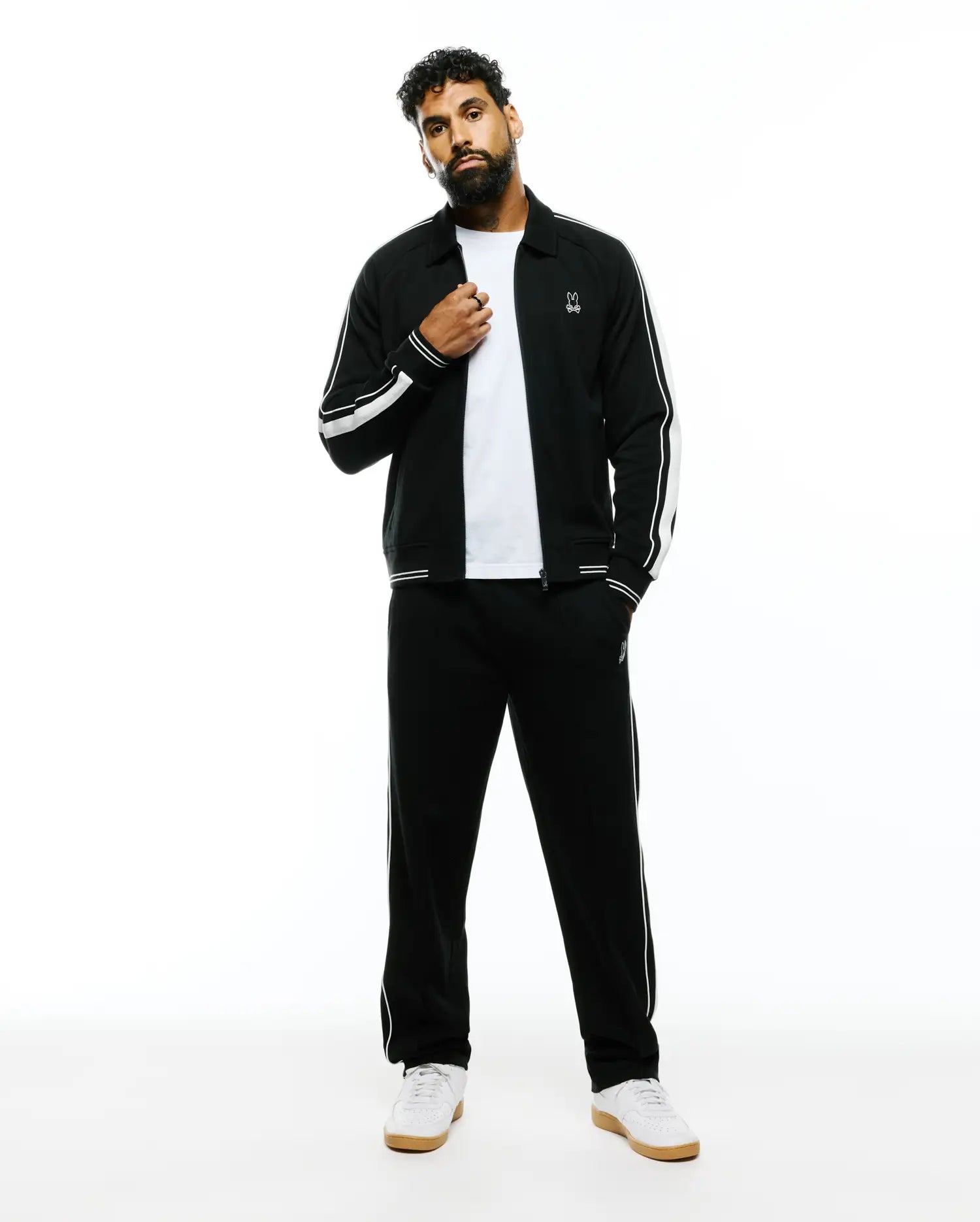 Black Mens Ceano Track Jacket and matching track pants, featuring white double sleeve stripes, single pant stripe, and small embroidered white Psycho Bunny skull logos on chest and thigh. Relaxed fit, smooth knit.