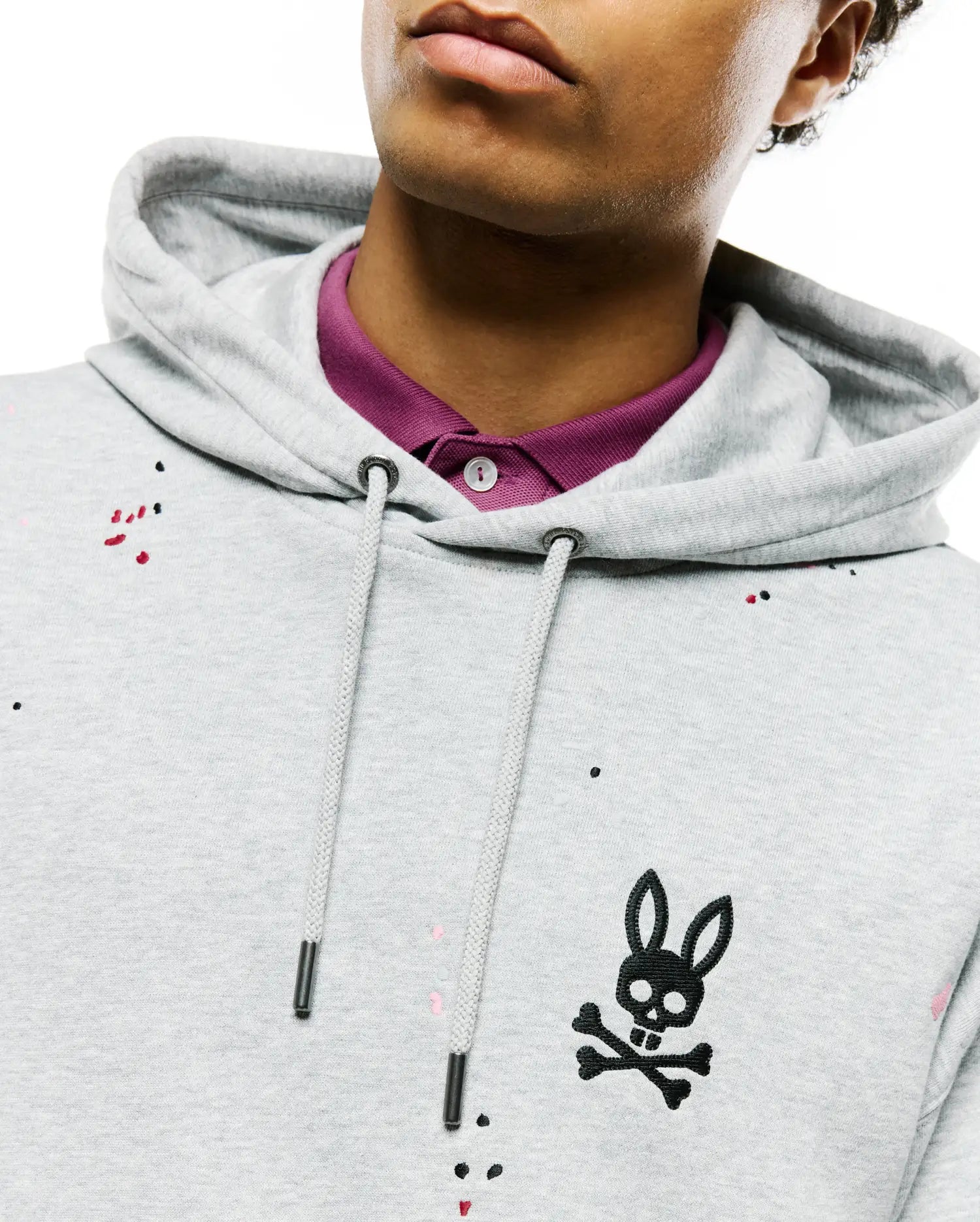 Mens Malcom Splatter Hoodie, a light grey french terry hoodie with a classic fit, featuring a black embroidered bunny skull logo on the left chest, drawstring hood, and colorful splatter paint accents.