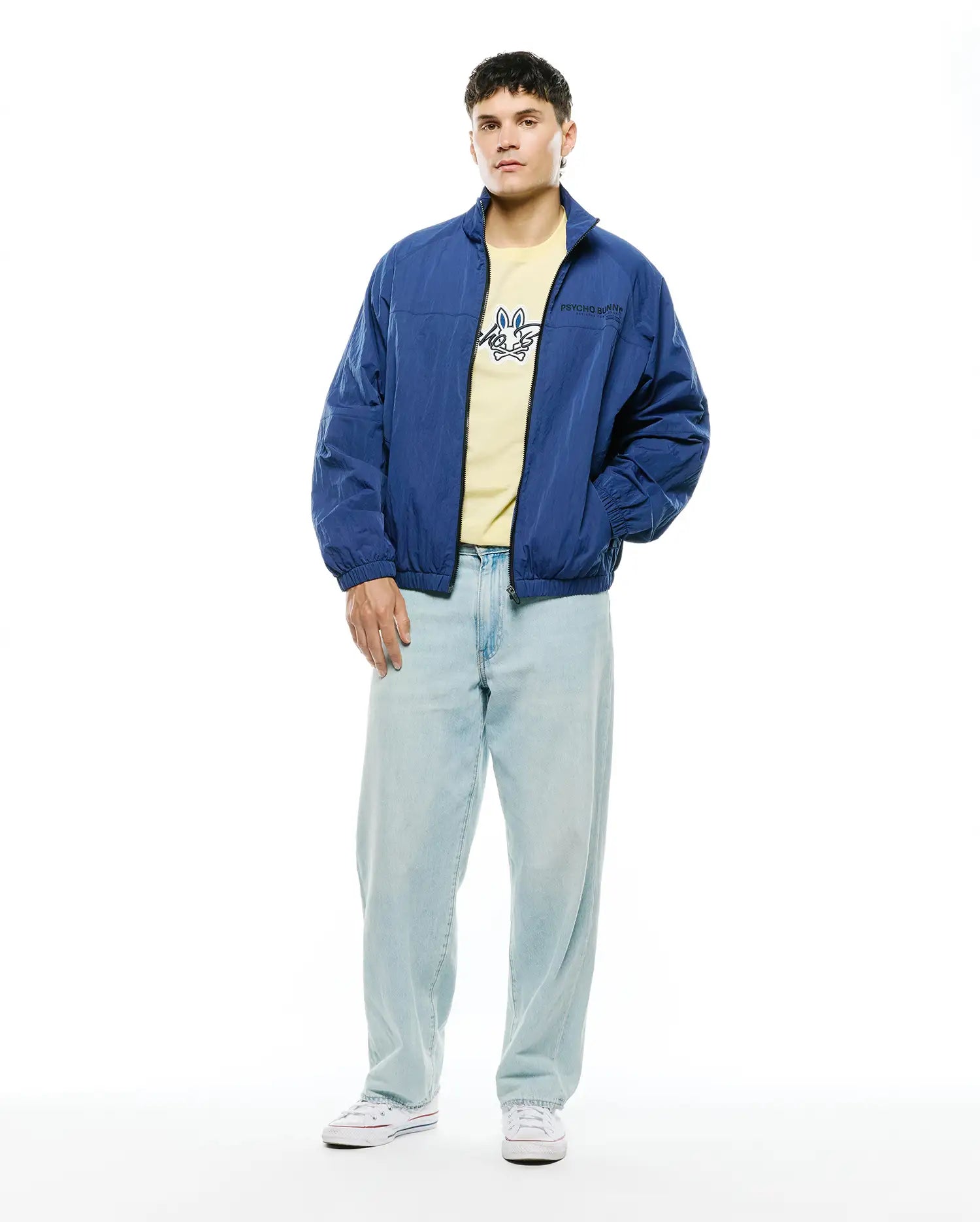 Mens Alder Nylon Jacket, a relaxed fit royal blue nylon windbreaker with a subtle Psycho Bunny chest logo, over a pale yellow jersey tee featuring a large blue bunny skull graphic, paired with light wash relaxed denim. (4)