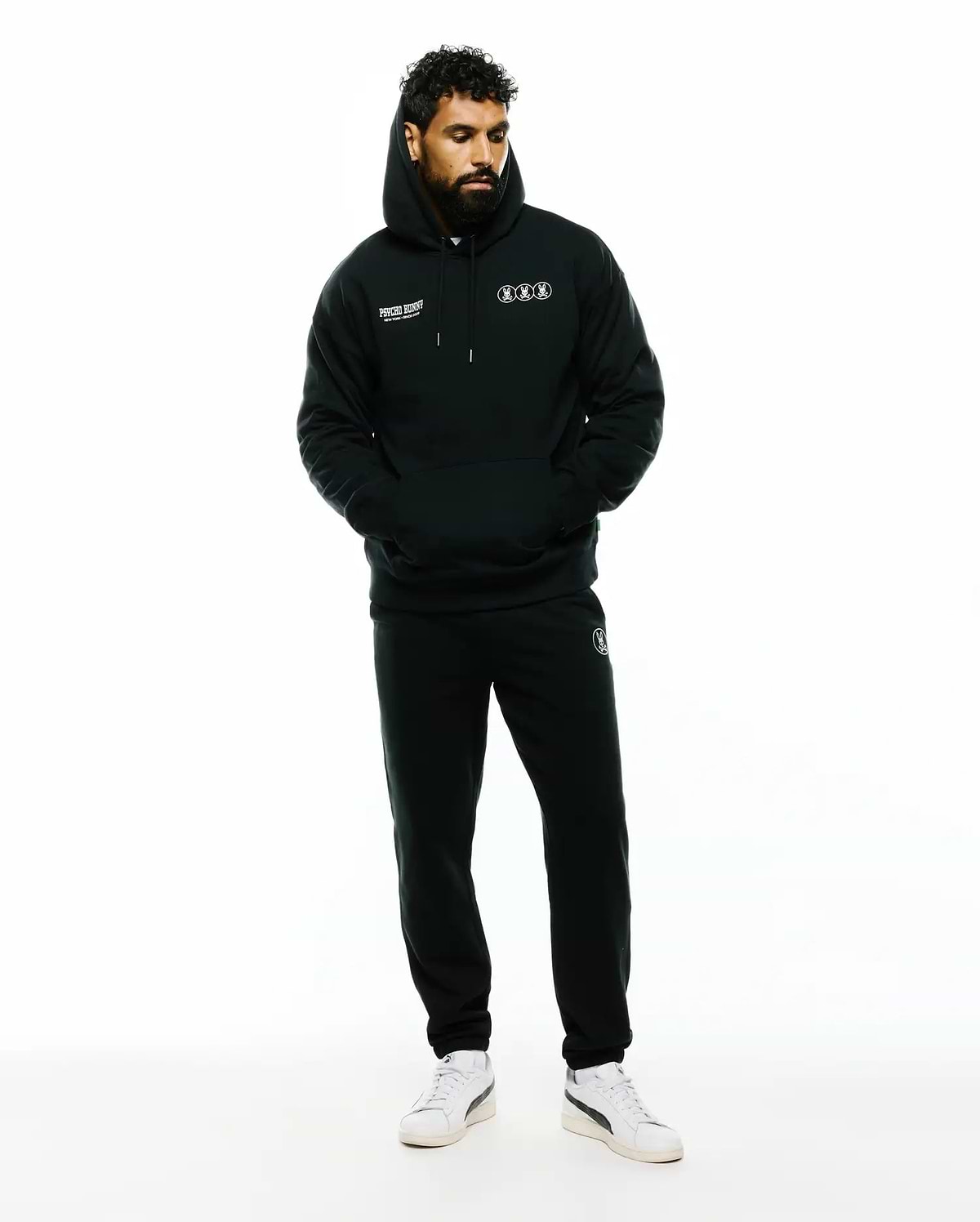 Mens Nathaniel Relaxed Chest Hoodie in black French terry, featuring white Psycho Bunny text and three outlined bunny skull logos on the chest, paired with matching black sweatpants with a small white bunny skull logo on the thigh. (2)