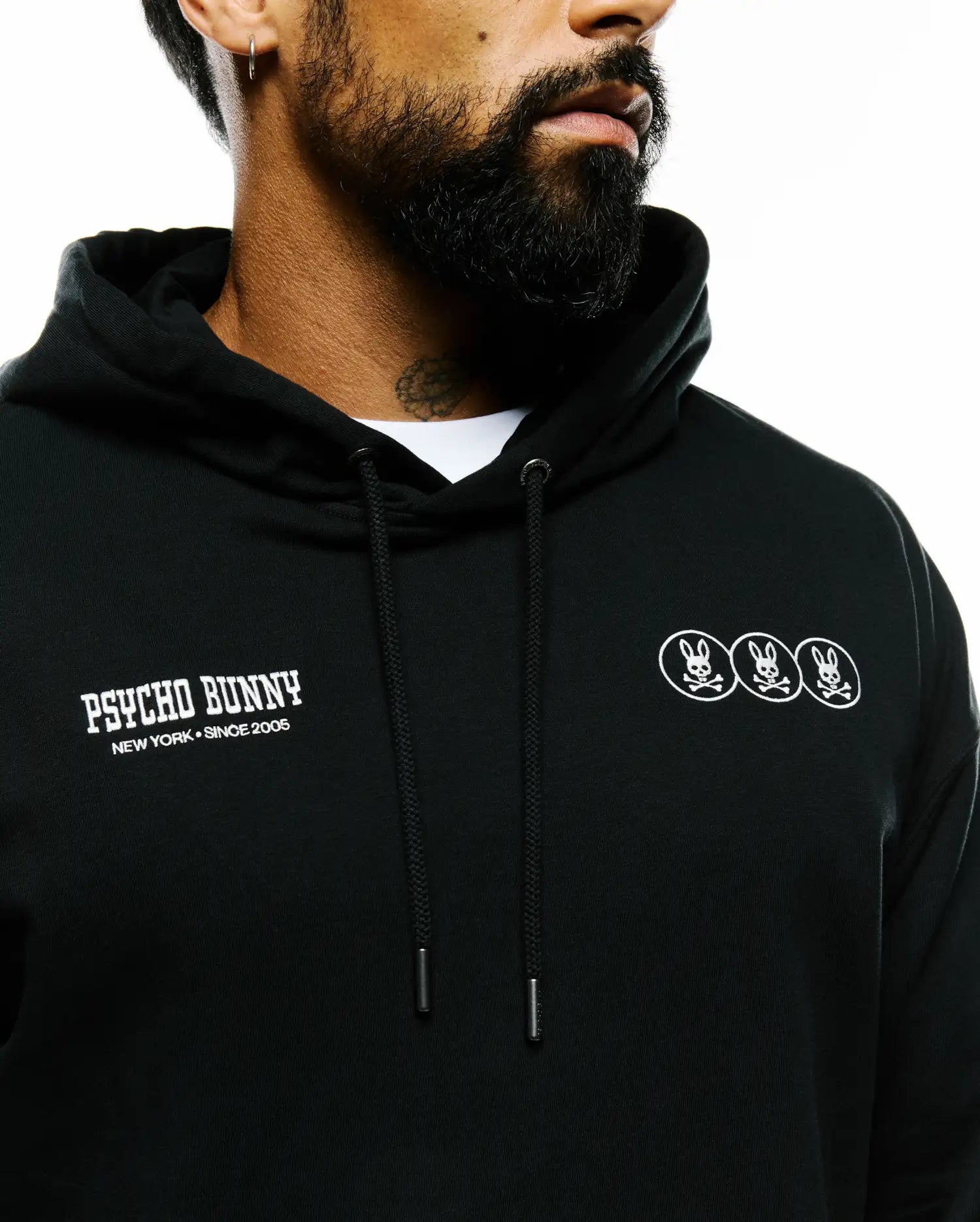 Mens Nathaniel Relaxed Chest Hoodie in black French terry, featuring white Psycho Bunny text and three bunny skull logos on the chest, with hood drawstrings. (4)