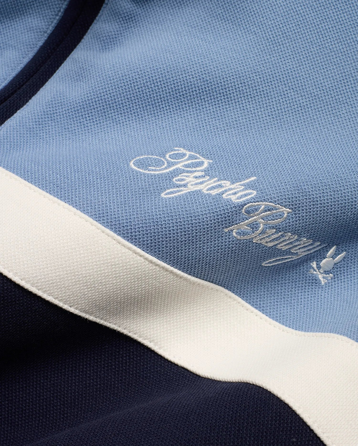 Men's Jacques Pique Track Jacket in light blue and navy pique fabric with a white stripe, featuring an embroidered white Psycho Bunny script logo and small bunny skull logo on the chest. (6)