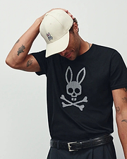 MENS STAN GRAPHIC TEE in black jersey, featuring a large white checkered Psycho Bunny skull and crossbones graphic on the chest. Accessorized with a cream cap with a small embroidered pink Psycho Bunny logo and a black leather belt.