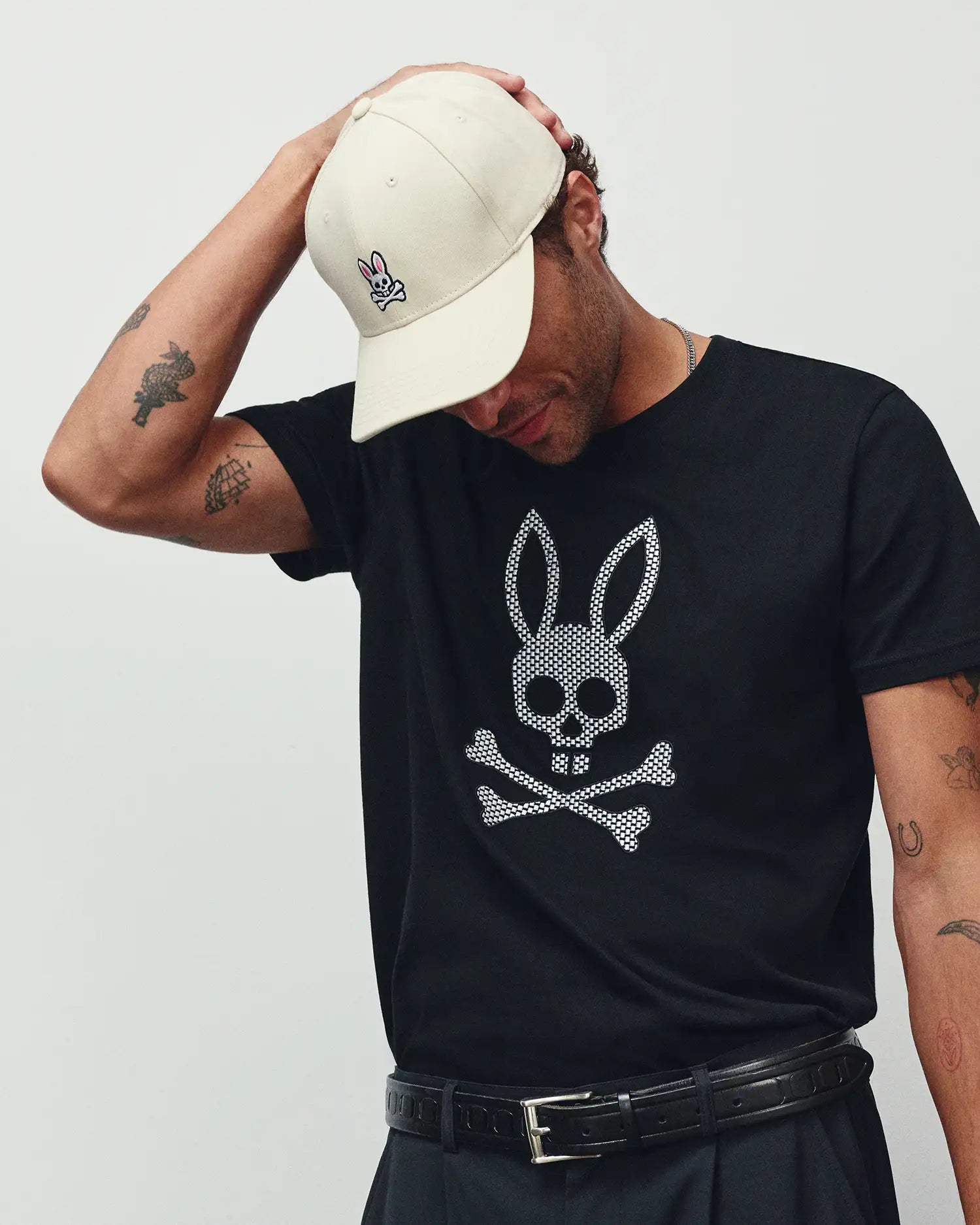 MENS STAN GRAPHIC TEE in black jersey, featuring a large white checkered Psycho Bunny skull and crossbones graphic on the chest. Accessorized with a cream cap with a small embroidered pink Psycho Bunny logo and a black leather belt.
