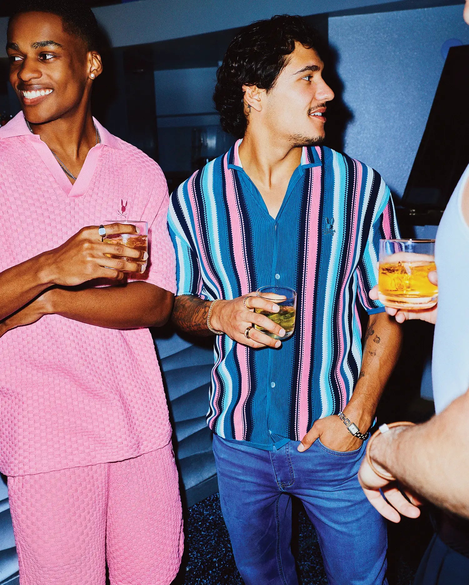 Mens Cliff Shirt: A relaxed fit, short-sleeve button-up with vibrant blue, pink, white, and black vertical stripes, featuring an embroidered Psycho Bunny logo. Also, a pink textured pique polo and shorts.