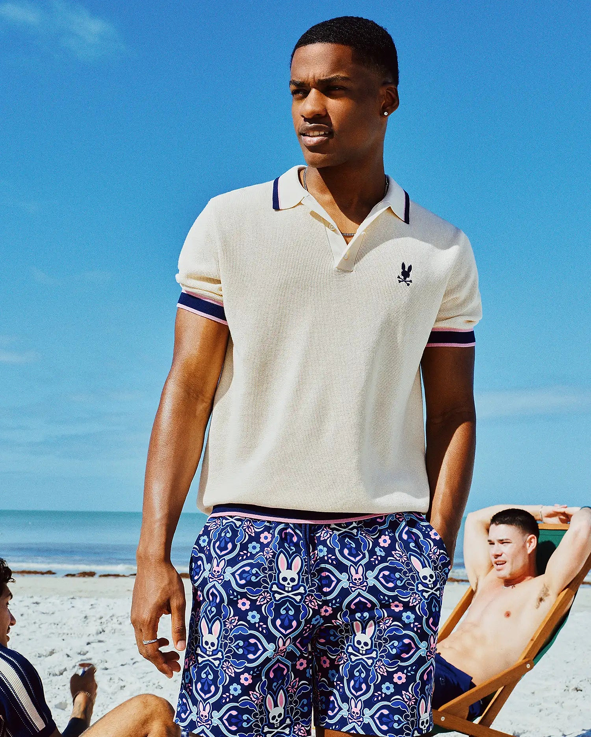Mens Dax Knit Polo, cream pique, with navy/pink striped collar/cuffs and small navy embroidered Psycho Bunny chest logo. Navy shorts feature an all-over pink/blue skull and floral print. (1)