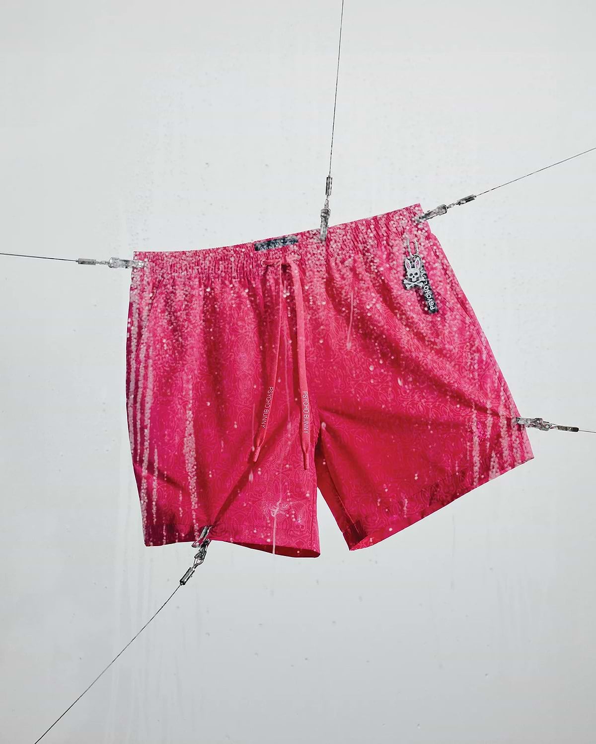 Mens Samos Hydrochromic Printed Swim Trunk 6, a hot pink relaxed fit swim trunk with a tonal pattern, white hydrochromic rain print, Psycho Bunny branded drawstrings, and a bunny skull logo charm. (2)