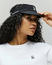 A woman with curly hair wears the Psycho Bunny WOMEN'S SPORT VISOR - WO1500001, black with an embroidered bunny logo and "Psycho Bunny" text, paired with a white tee featuring a small pink-and-black bunny logo on the chest.