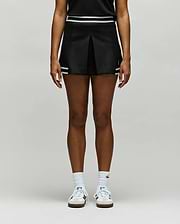 A person stands against a plain background, wearing the Psycho Bunny WOMENS KNIT SKORT - WO1900001 in black with white stripes, paired with a black top, white socks, and white sneakers. The image is cropped from the shoulders down. (2)
