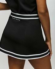 A person wears the Psycho Bunny WOMENS KNIT SKORT - WO1900001, a black skort with white stripes and "Psycho Bunny" embroidered on the waistband, standing with one hand resting at their side. (3)