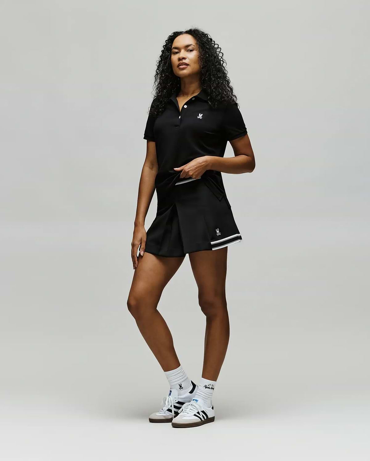 A confident woman with curly hair wears Psycho Bunny athletic wear: a black polo, WOMENS KNIT SKORT - WO1900001, white socks, and white sneakers, posing against a plain light background. (5)