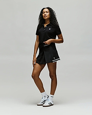 A confident woman with curly hair wears Psycho Bunny athletic wear: a black polo, WOMENS KNIT SKORT - WO1900001, white socks, and white sneakers, posing against a plain light background. (5)