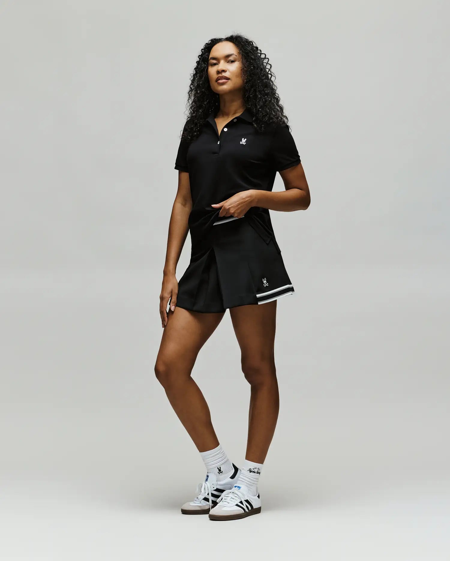 A confident woman with curly hair wears Psycho Bunny athletic wear: a black polo, WOMENS KNIT SKORT - WO1900001, white socks, and white sneakers, posing against a plain light background. (5)