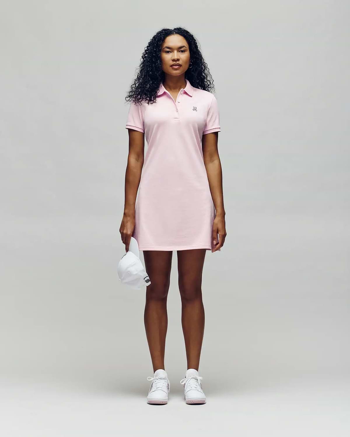 A person faces forward in a Psycho Bunny WOMENS POLO DRESS - WO2000003, a light pink polo dress with a feminine silhouette, styled with white sneakers and holding a white baseball cap against a plain light gray background. (4)