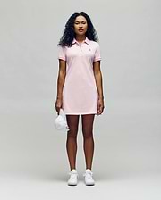 A person faces forward in a Psycho Bunny WOMENS POLO DRESS - WO2000003, a light pink polo dress with a feminine silhouette, styled with white sneakers and holding a white baseball cap against a plain light gray background. (4)