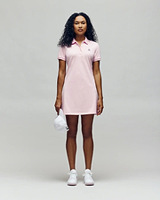 A person faces forward in a Psycho Bunny WOMENS POLO DRESS - WO2000003, a light pink polo dress with a feminine silhouette, styled with white sneakers and holding a white baseball cap against a plain light gray background. (4)