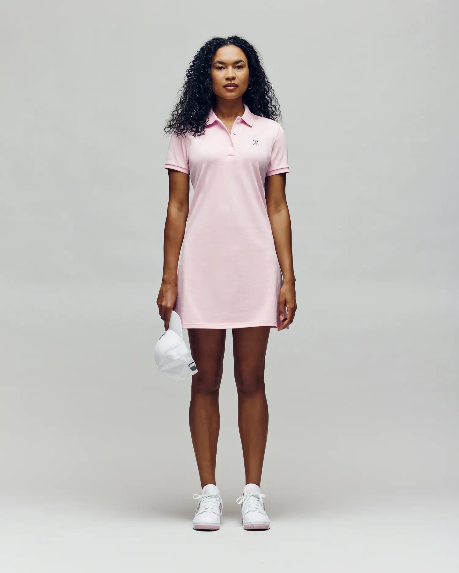 A person faces forward in a Psycho Bunny WOMENS POLO DRESS - WO2000003, a light pink polo dress with a feminine silhouette, styled with white sneakers and holding a white baseball cap against a plain light gray background. (4)