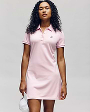 A woman with curly hair wears the Psycho Bunny WOMENS POLO DRESS - WO2000003 in light pink, featuring short sleeves, a collared neckline with buttons, and a small embroidered logo. She holds a white cap against a plain light background. (2)