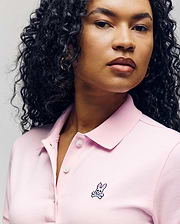A woman with curly hair wears the Psycho Bunny WOMENS POLO DRESS - WO2000003 in light pink, featuring a feminine silhouette, Mother-of-Pearl buttons, and an embroidered bunny and crossbones logo on the chest. (3)