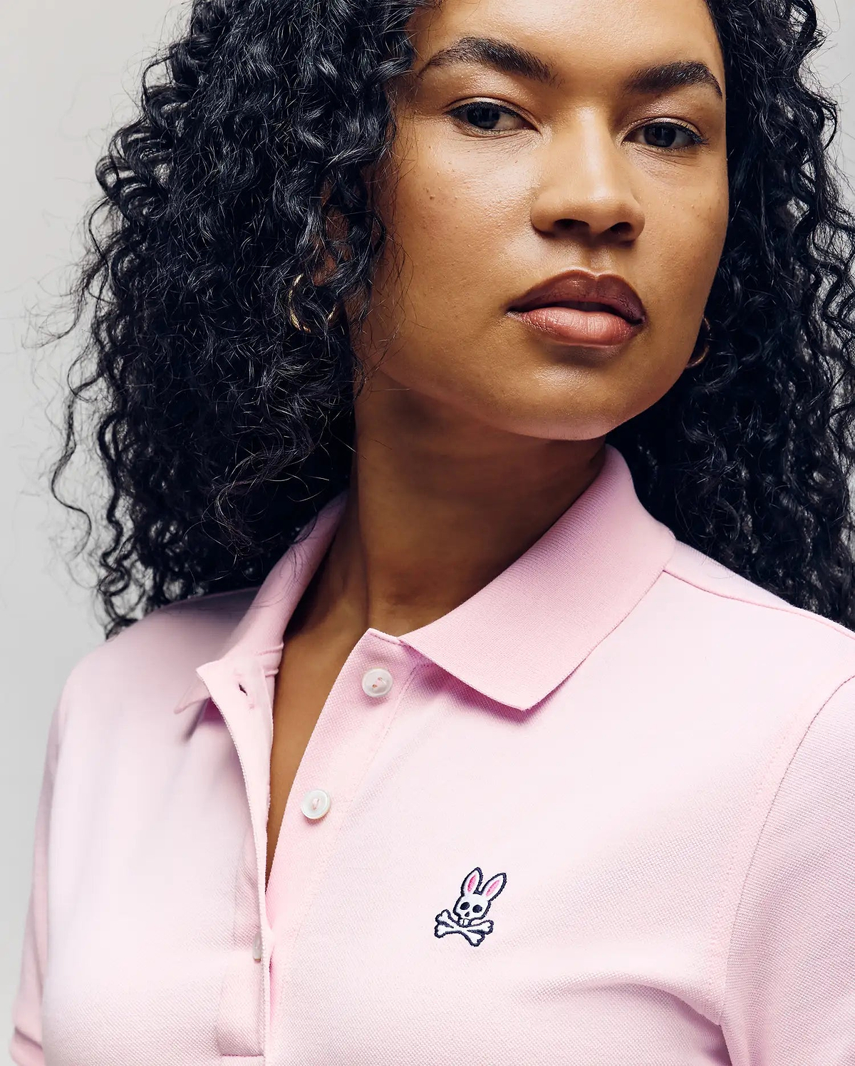 A woman with curly hair wears the Psycho Bunny WOMENS POLO DRESS - WO2000003 in light pink, featuring a feminine silhouette, Mother-of-Pearl buttons, and an embroidered bunny and crossbones logo on the chest. (3)