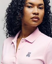 A woman with curly hair wears the Psycho Bunny WOMENS POLO DRESS - WO2000003 in light pink, featuring a feminine silhouette, Mother-of-Pearl buttons, and an embroidered bunny and crossbones logo on the chest. (3)