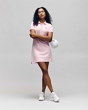 A confident person with curly hair wears the Psycho Bunny WOMENS POLO DRESS - WO2000003 in light pink, holding a white cap and pairing the look with classic white sneakers against a plain background. (1)