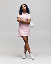 A confident person with curly hair wears the Psycho Bunny WOMENS POLO DRESS - WO2000003 in light pink, holding a white cap and pairing the look with classic white sneakers against a plain background. (1)