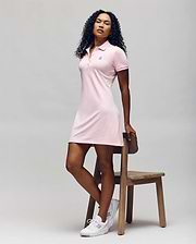 A woman with curly hair poses by a wooden chair, wearing the Psycho Bunny WOMENS POLO DRESS - WO2000003 in light pink with Mother-of-Pearl buttons and white sneakers, set against a plain light background. (5)