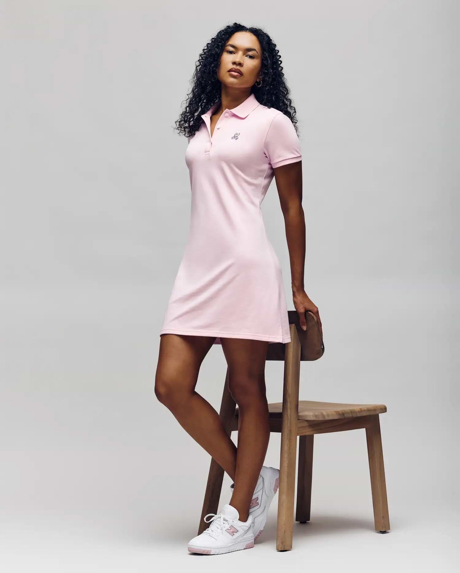 A woman with curly hair poses by a wooden chair, wearing the Psycho Bunny WOMENS POLO DRESS - WO2000003 in light pink with Mother-of-Pearl buttons and white sneakers, set against a plain light background. (5)