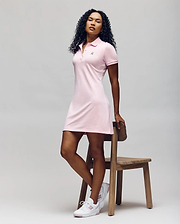 A woman with curly hair poses by a wooden chair, wearing the Psycho Bunny WOMENS POLO DRESS - WO2000003 in light pink with Mother-of-Pearl buttons and white sneakers, set against a plain light background. (5)