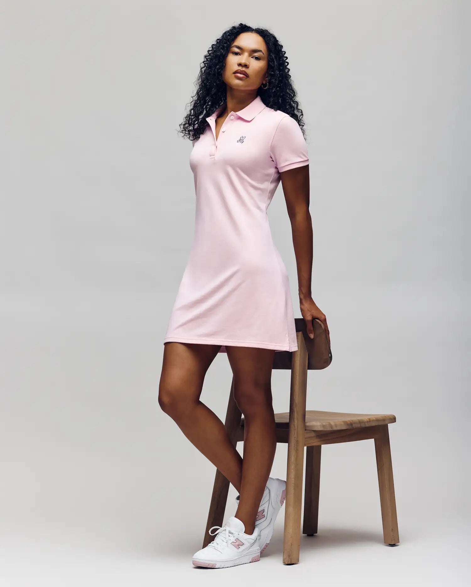 A woman with curly hair poses by a wooden chair, wearing the Psycho Bunny WOMENS POLO DRESS - WO2000003 in light pink with Mother-of-Pearl buttons and white sneakers, set against a plain light background. (5)
