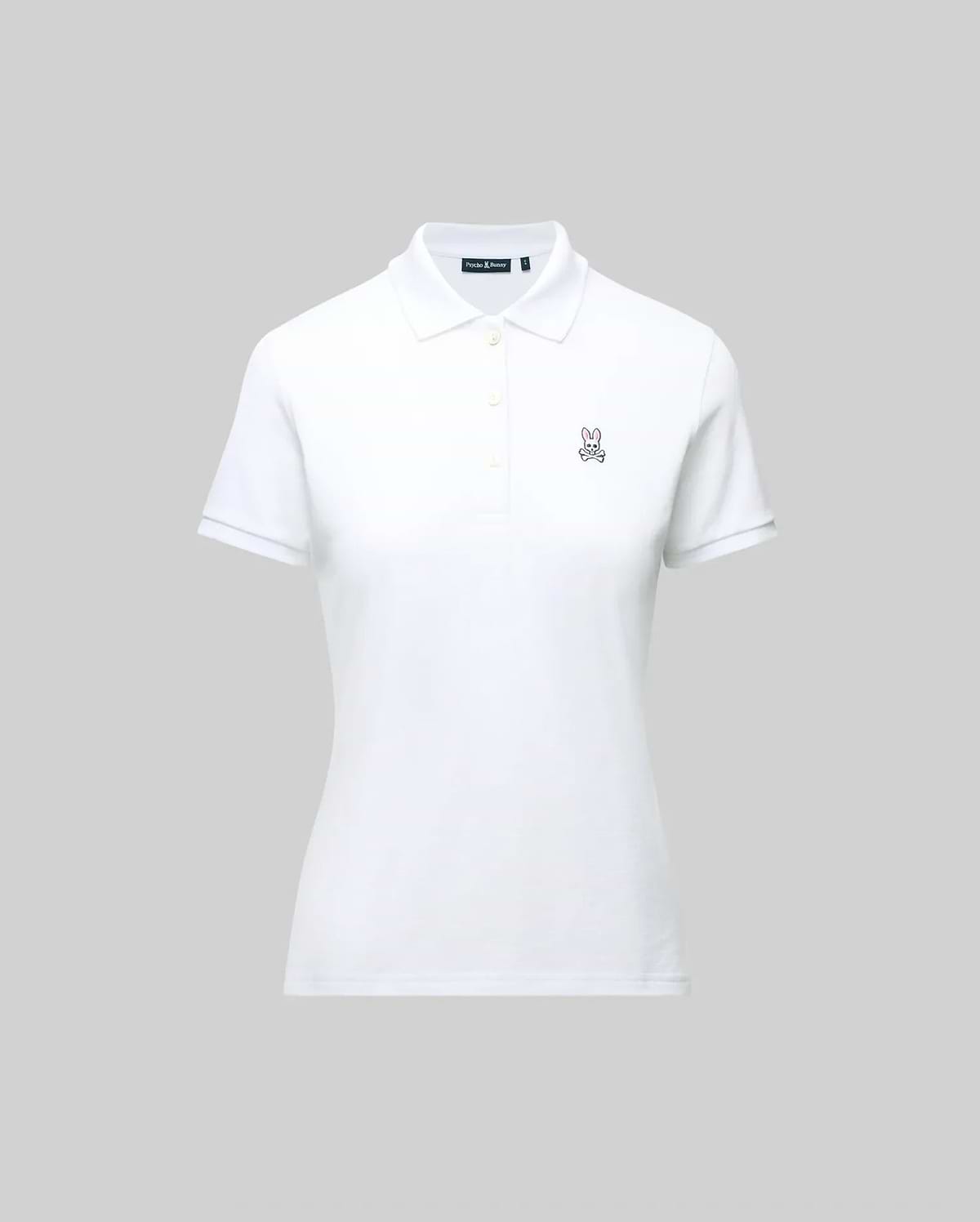 The WOMENS PIQUE POLO - WT0100003 by Psycho Bunny is a white pique polo featuring a collar, buttons, and the embroidered Bunny logo on the left chest. Crafted for Pima flexibility, it is presented against a plain grey background.