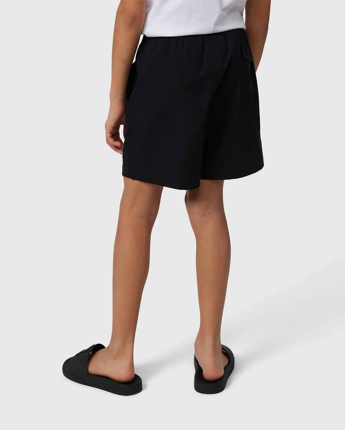 A person wearing a white T-shirt with a subtle bunny pattern stands facing away, showcasing the KIDS MALTA HYDROCHROMIC SWIM TRUNK in black by Psycho Bunny and black slip-on shoes against a neutral background. Their hands rest loosely at their sides.