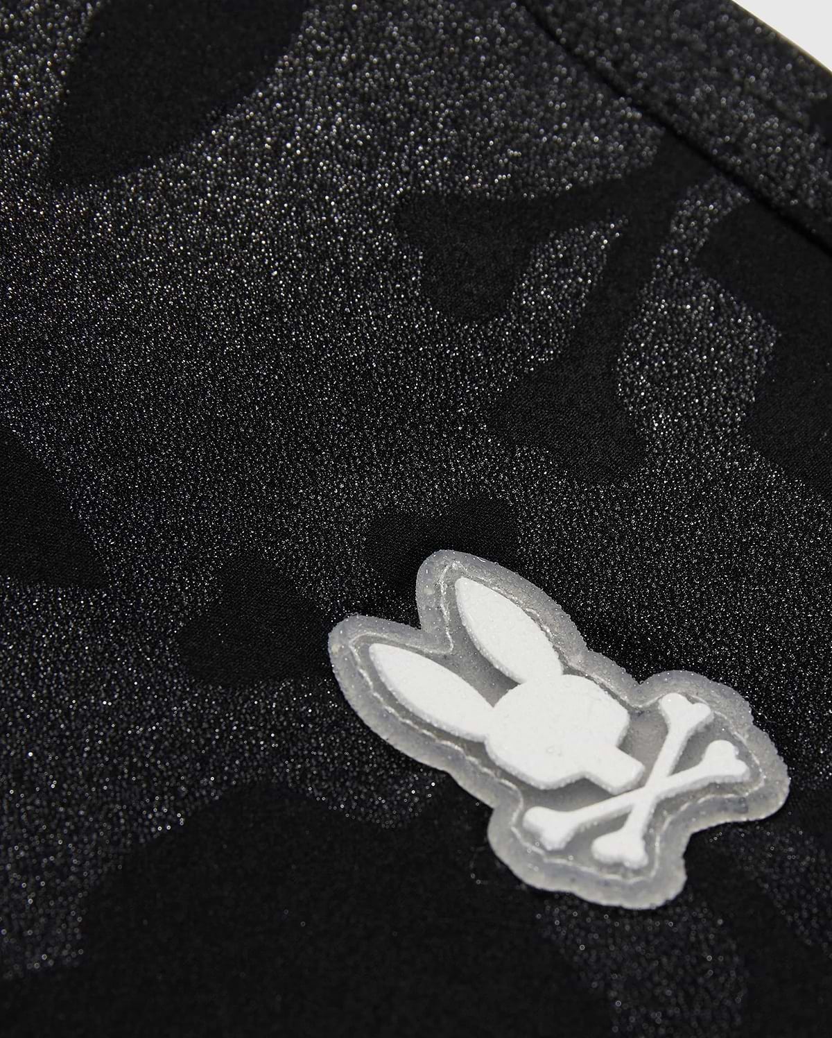 The KIDS MALTA HYDROCHROMIC SWIM TRUNK - B0W321B2SW by Psycho Bunny showcases a close-up of textured black fabric with a subtle Bunny pattern. A small, white, cartoon-style emblem displays a rabbit head with crossed bones beneath it, resembling a playful take on the skull and crossbones design—perfect for stylish swim trunks.