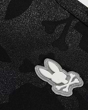 The KIDS MALTA HYDROCHROMIC SWIM TRUNK - B0W321B2SW by Psycho Bunny showcases a close-up of textured black fabric with a subtle Bunny pattern. A small, white, cartoon-style emblem displays a rabbit head with crossed bones beneath it, resembling a playful take on the skull and crossbones design—perfect for stylish swim trunks.