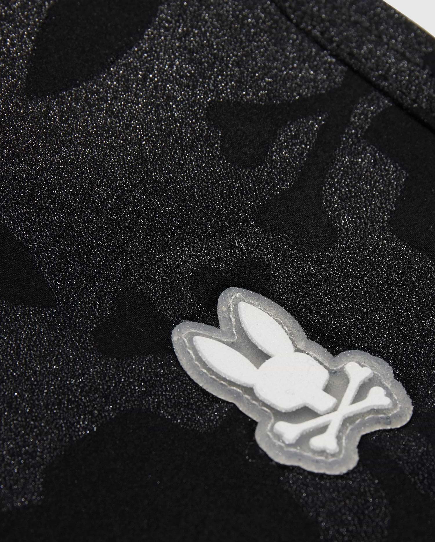 The KIDS MALTA HYDROCHROMIC SWIM TRUNK - B0W321B2SW by Psycho Bunny showcases a close-up of textured black fabric with a subtle Bunny pattern. A small, white, cartoon-style emblem displays a rabbit head with crossed bones beneath it, resembling a playful take on the skull and crossbones design—perfect for stylish swim trunks.