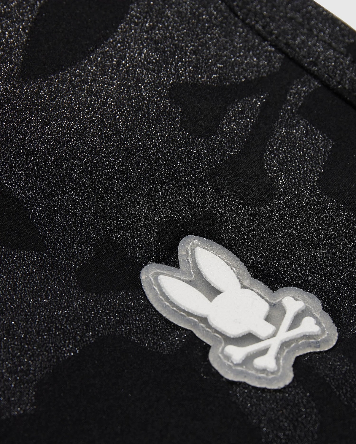 The KIDS MALTA HYDROCHROMIC SWIM TRUNK - B0W321B2SW by Psycho Bunny showcases a close-up of textured black fabric with a subtle Bunny pattern. A small, white, cartoon-style emblem displays a rabbit head with crossed bones beneath it, resembling a playful take on the skull and crossbones design—perfect for stylish swim trunks.