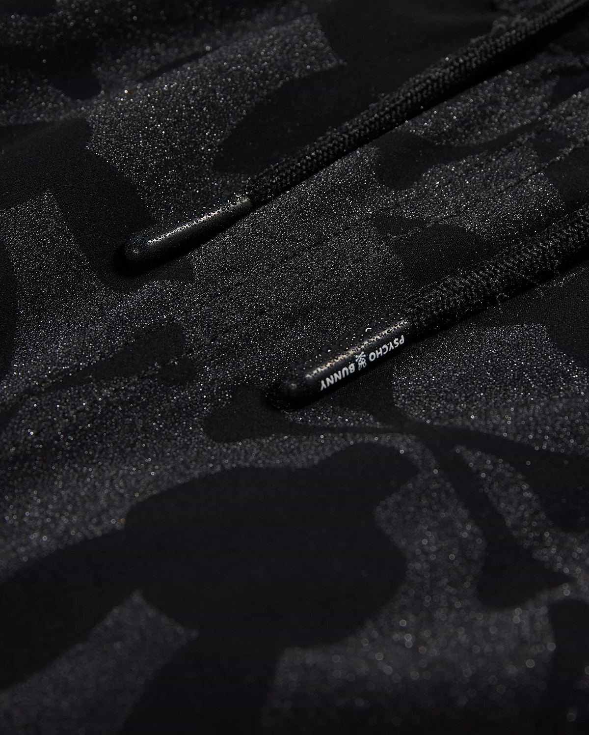 Close-up of Psycho Bunny's KIDS MALTA HYDROCHROMIC SWIM TRUNK - B0W321B2SW, showcasing a black textured fabric with a subtle camouflage pattern in shades of dark grey and black. Two parallel black cords with small text on one cord are partially visible, reminiscent of sleek swim trunks. The overall appearance is both modern and hydrochromic.