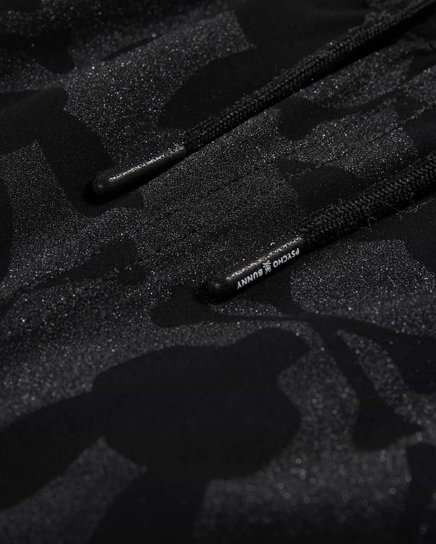 Close-up of Psycho Bunny's KIDS MALTA HYDROCHROMIC SWIM TRUNK - B0W321B2SW, showcasing a black textured fabric with a subtle camouflage pattern in shades of dark grey and black. Two parallel black cords with small text on one cord are partially visible, reminiscent of sleek swim trunks. The overall appearance is both modern and hydrochromic.