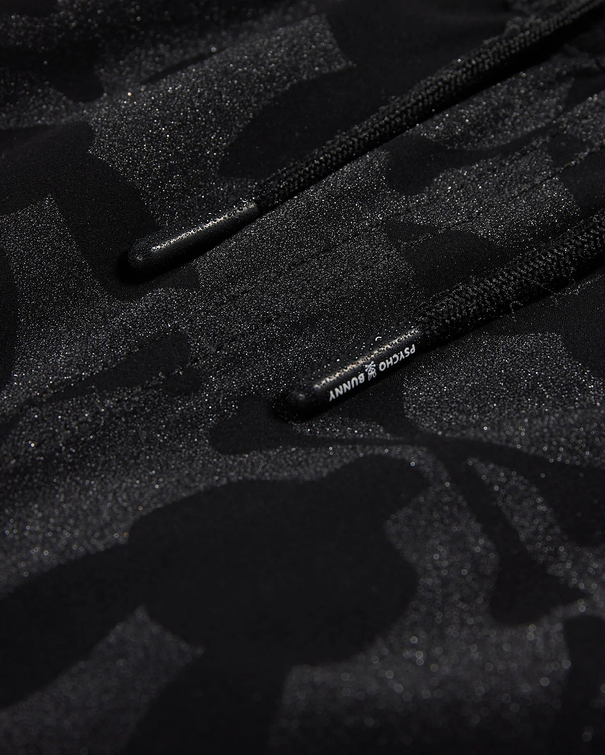 Close-up of Psycho Bunny's KIDS MALTA HYDROCHROMIC SWIM TRUNK - B0W321B2SW, showcasing a black textured fabric with a subtle camouflage pattern in shades of dark grey and black. Two parallel black cords with small text on one cord are partially visible, reminiscent of sleek swim trunks. The overall appearance is both modern and hydrochromic.